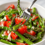 A bowl of fresh side dish topped with crumbled cheese, with a fork picking up some asparagus.