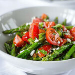A bowl of tomato and asparagus salad with feta cheese and seasonings, on a white tablecloth.