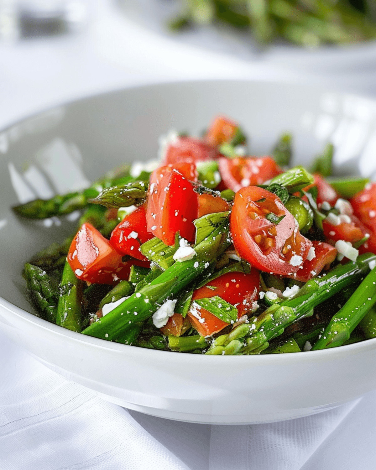 A bowl of tomato and asparagus salad with feta cheese and seasonings, on a white tablecloth.