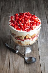 A strawberry angel food cake trifle in a glass bowl, with layers of whipped cream, sponge cake, and fresh strawberries, placed on a wooden surface with a spoon beside it.