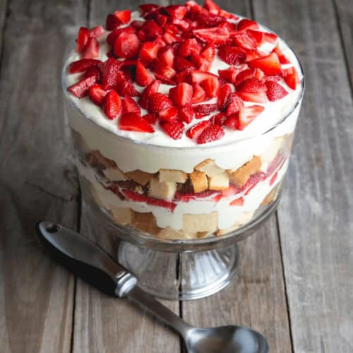 A strawberry angel food cake trifle in a glass bowl, with layers of whipped cream, sponge cake, and fresh strawberries, placed on a wooden surface with a spoon beside it.