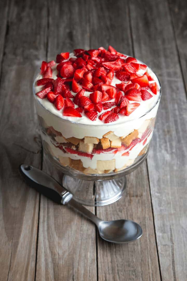 A strawberry angel food cake trifle in a glass bowl, with layers of whipped cream, sponge cake, and fresh strawberries, placed on a wooden surface with a spoon beside it.