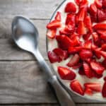 A bowl of sliced strawberries atop a creamy strawberry angel food cake dessert, paired with a stainless steel spoon on a wooden table.