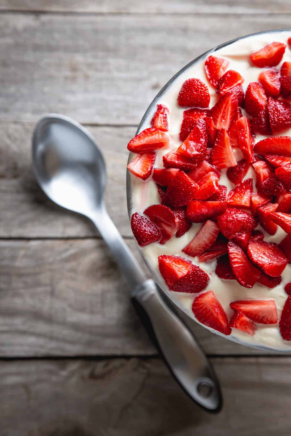 A bowl of sliced strawberries atop a creamy strawberry angel food cake dessert, paired with a stainless steel spoon on a wooden table.