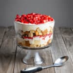 A layered trifle dessert in a glass bowl filled with strawberries, cream, and pieces of a strawberry angel food cake on a wooden surface, with a metal spoon beside it.