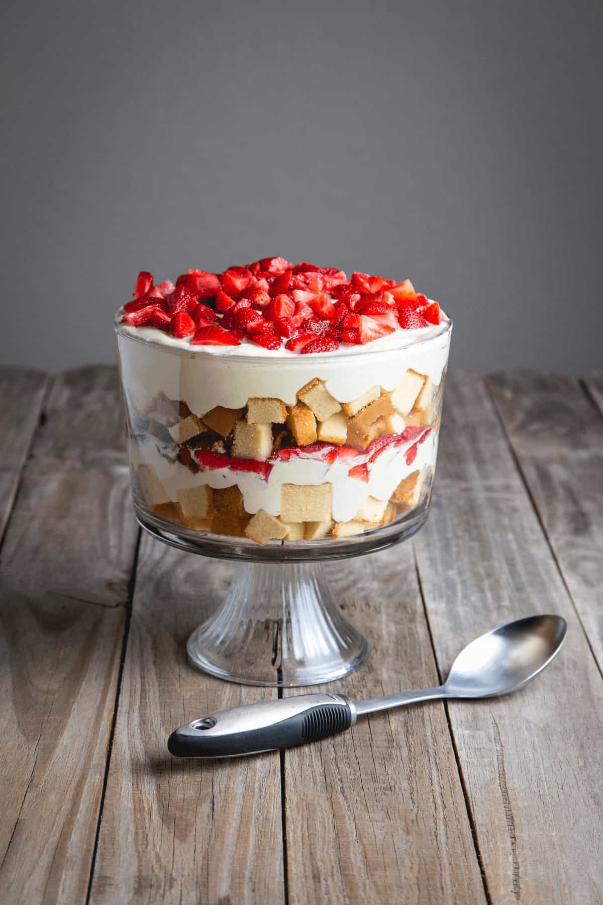 A layered trifle dessert in a glass bowl filled with strawberries, cream, and pieces of a strawberry angel food cake on a wooden surface, with a metal spoon beside it.