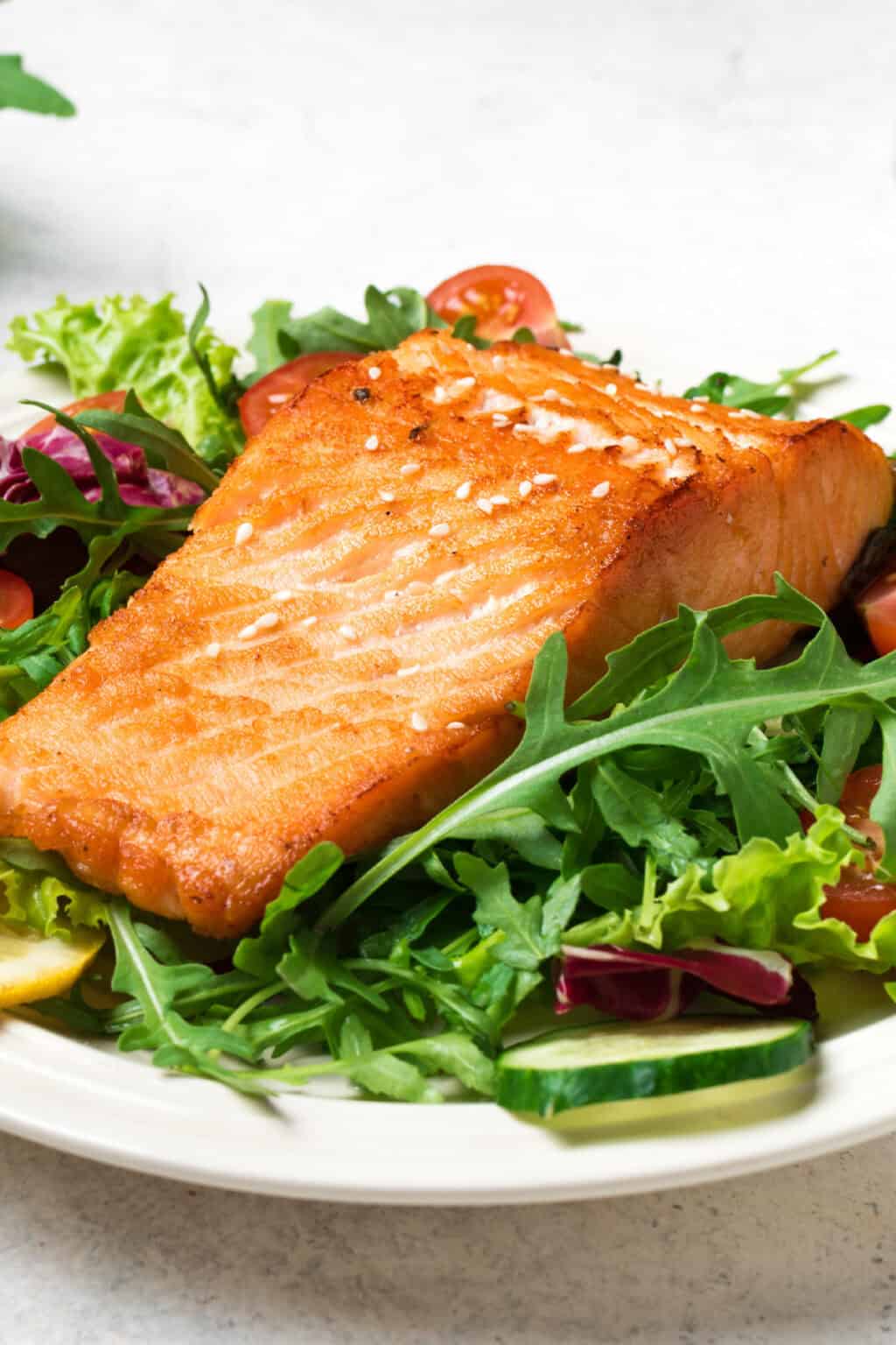 Grilled Salmon Salad: A succulent grilled salmon fillet rests elegantly on a vibrant bed of mixed greens, adorned with crisp sliced cucumbers and sweet cherry tomatoes, all artfully arranged on a pristine white plate.