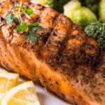 Grilled salmon fillet, artfully presented as a salad, garnished with herbs and served with fresh lemon wedges and a medley of steamed vegetables including broccoli.