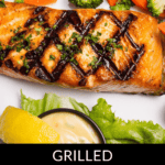 Grilled salmon salad features perfectly cooked salmon topped with fresh herbs, nestled among broccoli and carrots. A lemon wedge adds zest, while a tasty sauce on the side completes this delightful dish.