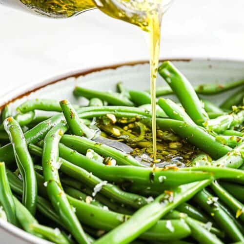 A hand pours olive oil and herb dressing from a jar onto a plate of cooked green beans, creating a vibrant Green Bean Salad.