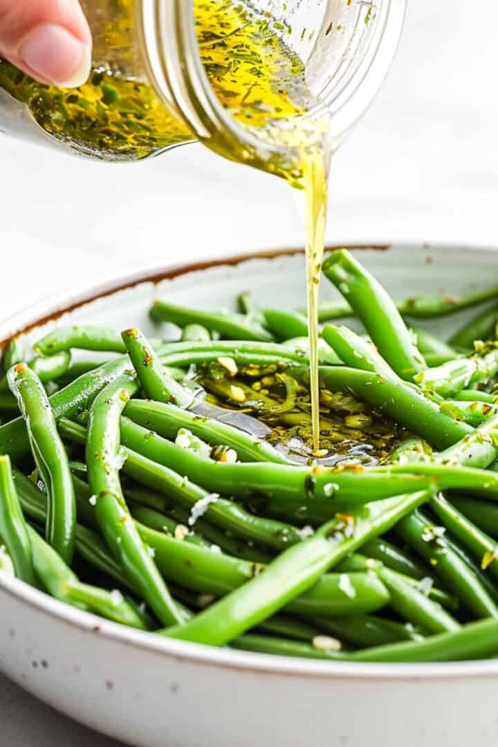 A hand pours olive oil and herb dressing from a jar onto a plate of cooked green beans, creating a vibrant Green Bean Salad.