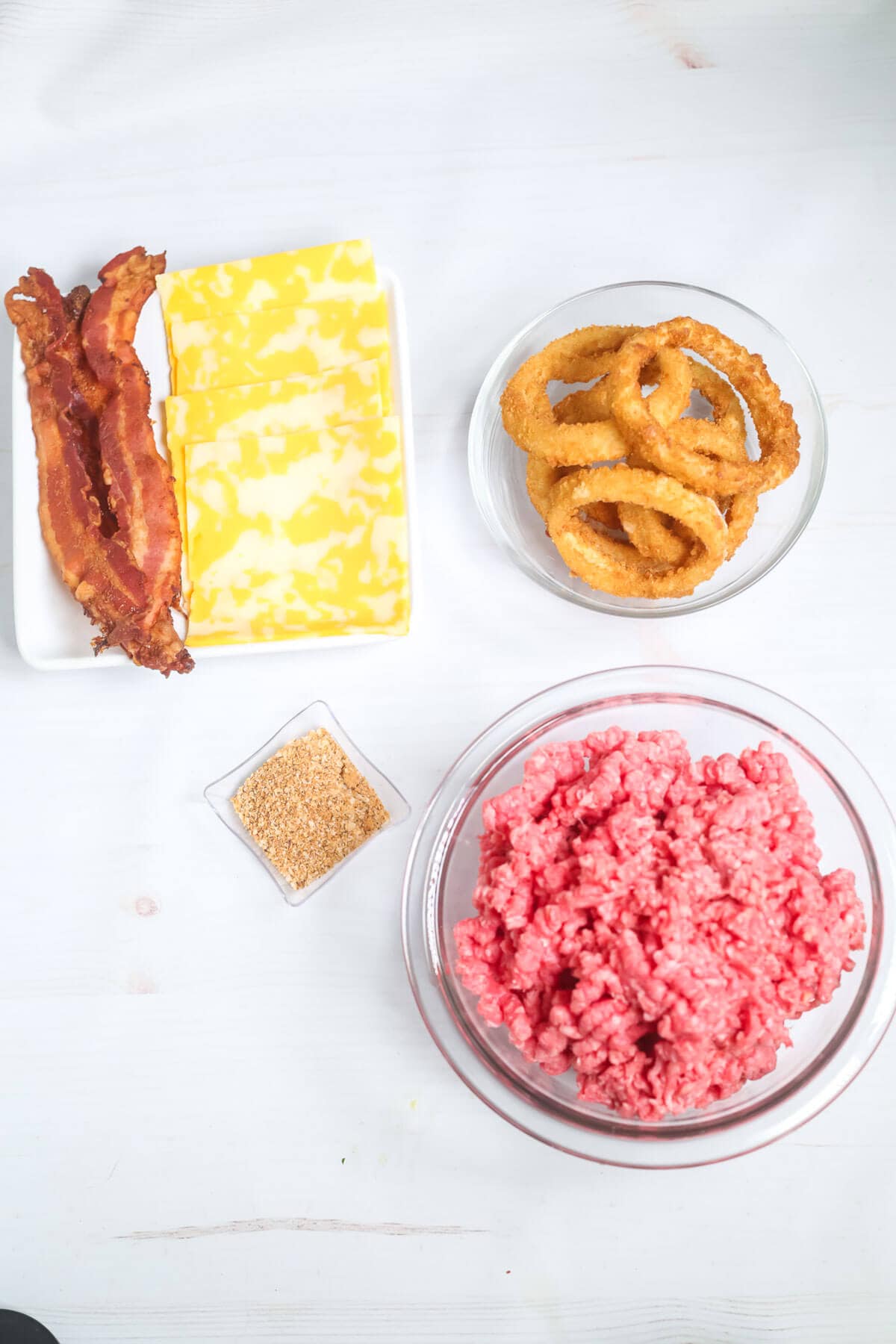 A white surface displaying raw ground beef, bacon strips, cheese slices, onion rings, and a small dish of seasoning—everything you need for the ultimate Onion Ring Burger.