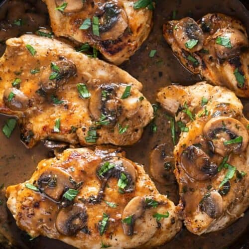 Four cooked chicken breasts in a skillet topped with sautéed mushrooms and garnished with chopped parsley in a rich Marsala sauce, perfect for serving over fettuccine for an authentic Fettuccine Chicken Marsala experience.