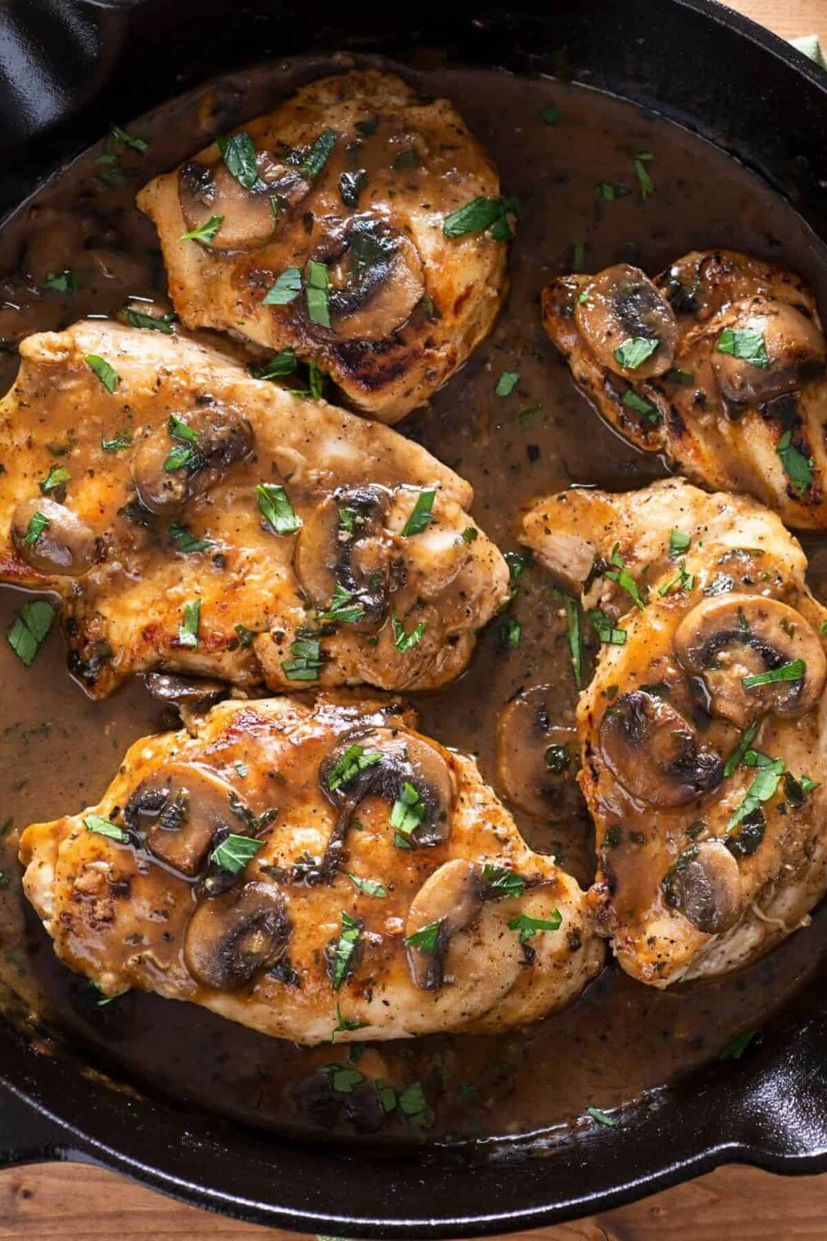 Four cooked chicken breasts in a skillet topped with sautéed mushrooms and garnished with chopped parsley in a rich Marsala sauce, perfect for serving over fettuccine for an authentic Fettuccine Chicken Marsala experience.