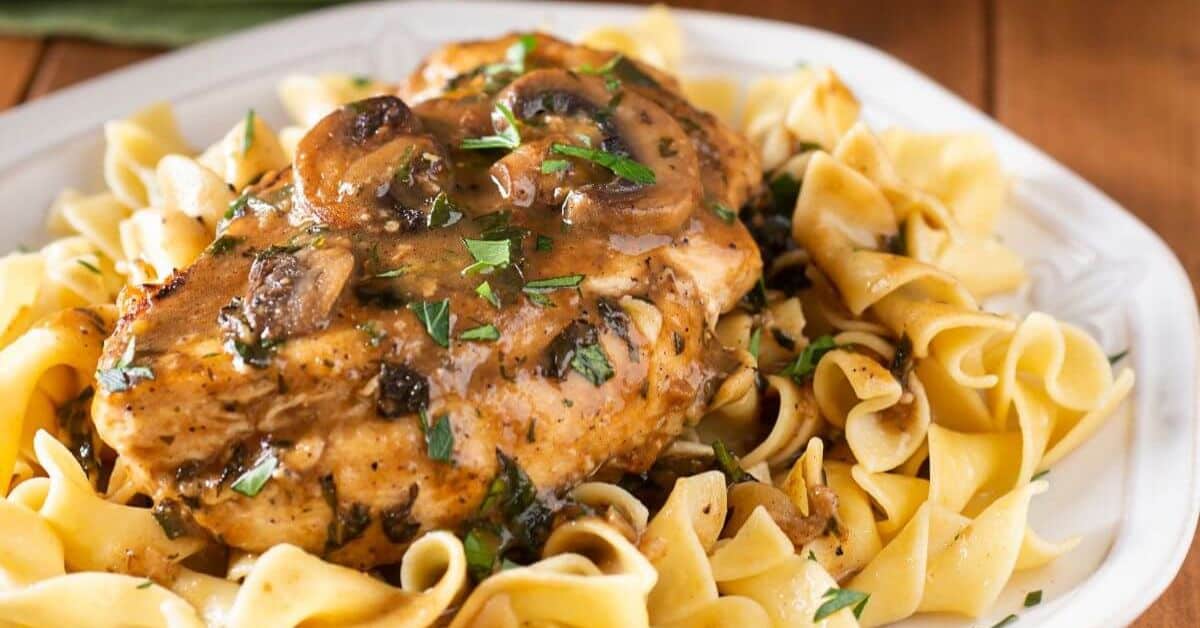 Fettuccine Chicken Marsala features tender chicken breast with mushroom gravy over egg noodles, garnished with chopped parsley on a white plate.
