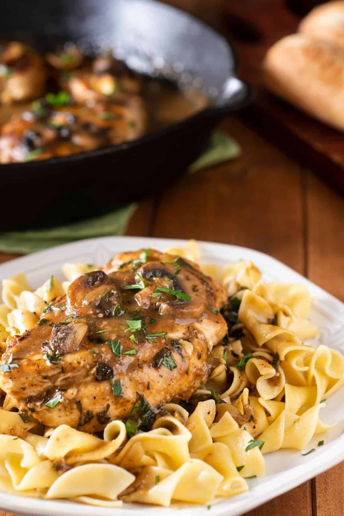 A plate of meat topped with mushroom gravy and garnished with herbs sits in front of a cast-iron skillet on a wooden table.