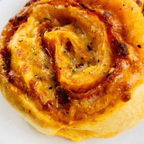 A close-up of a golden-brown pizza roll up, showcasing layers of baked dough with visible herbs and melted cheese, rests invitingly on a white plate.