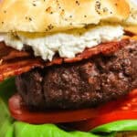 A close-up of a Steakhouse Burger with a beef patty, bacon, lettuce, tomato, and sauce, all served in a seeded bun. A knife is stuck into the top of the burger.