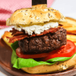 A close-up of a gourmet Sandwich with lettuce, tomato, bacon, and a beef patty, topped with a bun through which a wooden-handled knife is inserted. A side of fries is visible in the background.