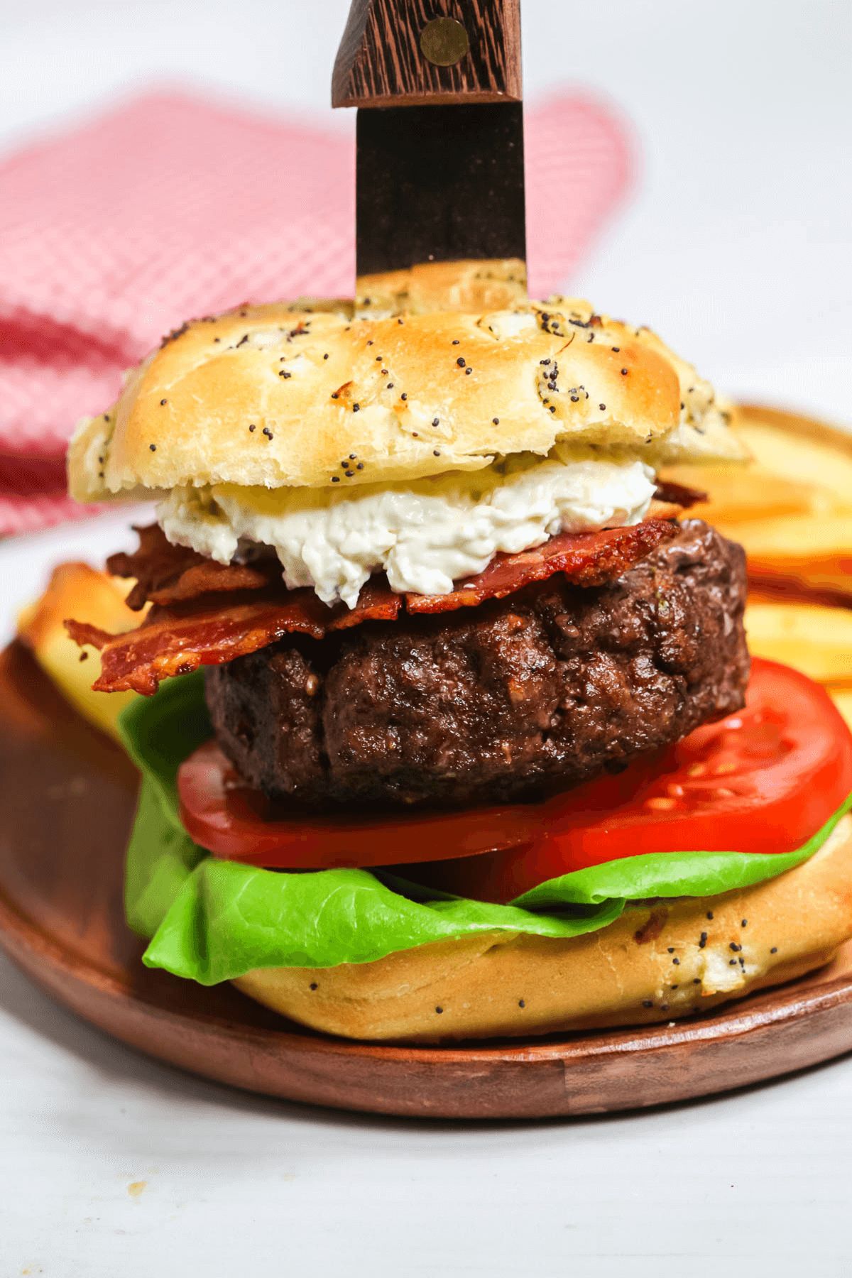 A close-up of a gourmet Sandwich with lettuce, tomato, bacon, and a beef patty, topped with a bun through which a wooden-handled knife is inserted. A side of fries is visible in the background.