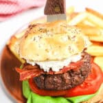 A close-up of a decadent Steakhouse Burger with a knife stabbed through the top bun. The burger contains lettuce, tomato, bacon, and cheese, and is accompanied by a side of fries on a wooden plate.