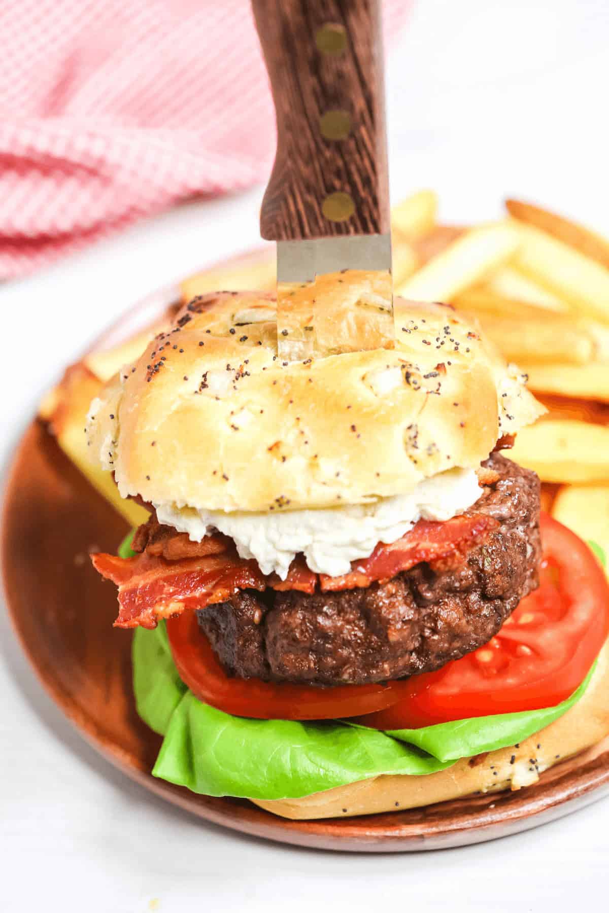A close-up of a decadent Steakhouse Burger with a knife stabbed through the top bun. The burger contains lettuce, tomato, bacon, and cheese, and is accompanied by a side of fries on a wooden plate.