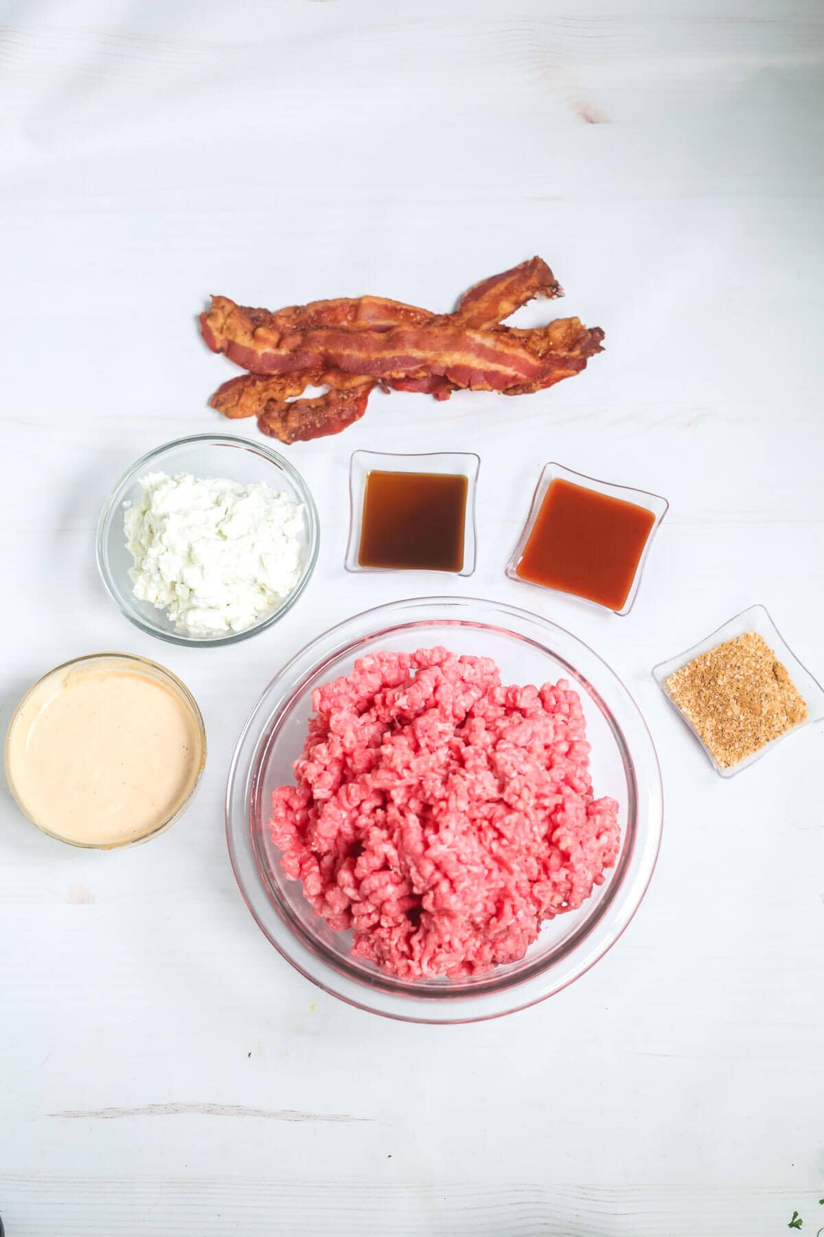 Overhead view of a bowl filled with raw ground beef, surrounded by dishes of white sauce, red sauce, brown sauce, seasoning, cooked bacon strips, and a bowl of minced white vegetables—perfect ingredients to craft a delightful Burger.