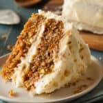 A slice of pumpkin carrot cake with cream cheese frosting and walnut pieces sits on a white plate. A larger piece of the same cake is in the background.