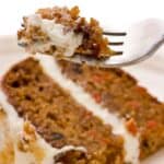 A fork holding a bite of pumpkin carrot cake with cream cheese frosting is positioned above a slice of the cake on a white plate.