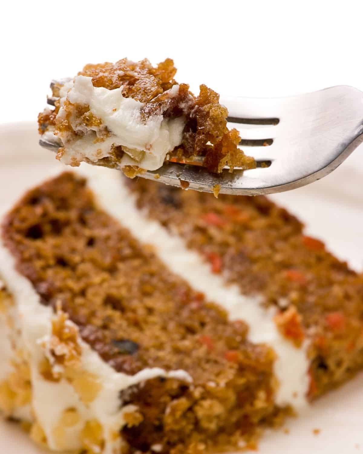 A fork holding a bite of pumpkin carrot cake with cream cheese frosting is positioned above a slice of the cake on a white plate.