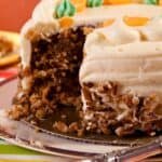 A sliced pumpkin carrot cake with cream cheese frosting and pecan decoration sits on a silver serving plate, with one piece missing and a portion on a small plate in the background.