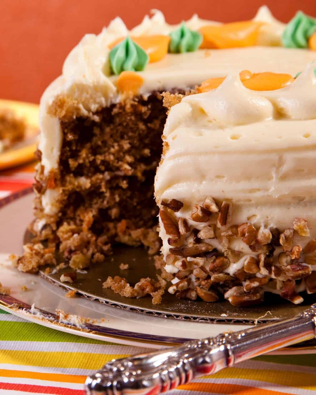 A sliced pumpkin carrot cake with cream cheese frosting and pecan decoration sits on a silver serving plate, with one piece missing and a portion on a small plate in the background.