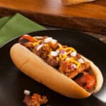 A chili cheese dog in a bun is topped with chili, shredded cheese, and chopped onions, served on a black plate with a green napkin.