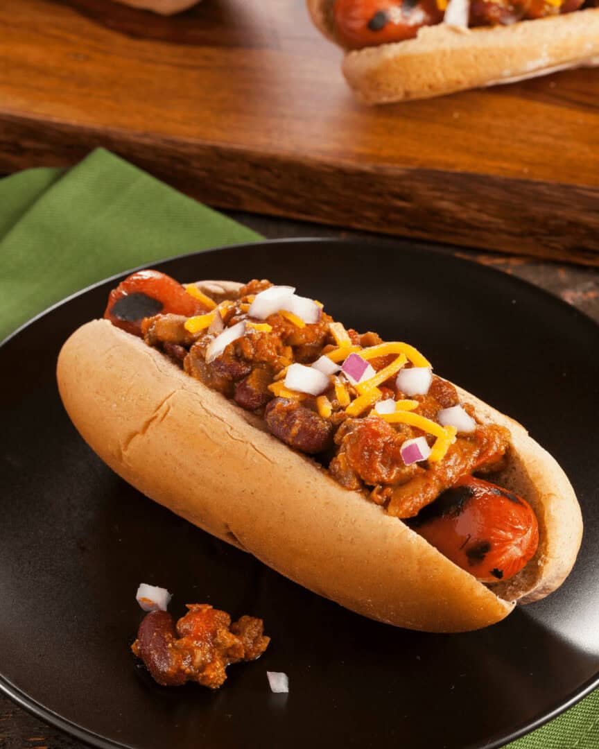 A chili cheese dog in a bun is topped with chili, shredded cheese, and chopped onions, served on a black plate with a green napkin.