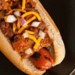 A chili cheese dog in a bun, topped with shredded cheese and finely chopped onions, sits on a black plate.