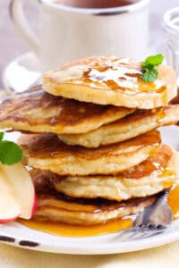 A stack of whole wheat apple cinnamon pancakes drizzled with syrup and garnished with mint sits invitingly on a plate, accompanied by fresh apple slices. A steaming cup of coffee completes the cozy breakfast scene in the background.