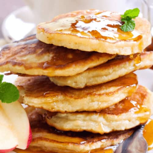 A stack of whole wheat apple cinnamon pancakes drizzled with syrup and garnished with mint sits invitingly on a plate, accompanied by fresh apple slices. A steaming cup of coffee completes the cozy breakfast scene in the background.