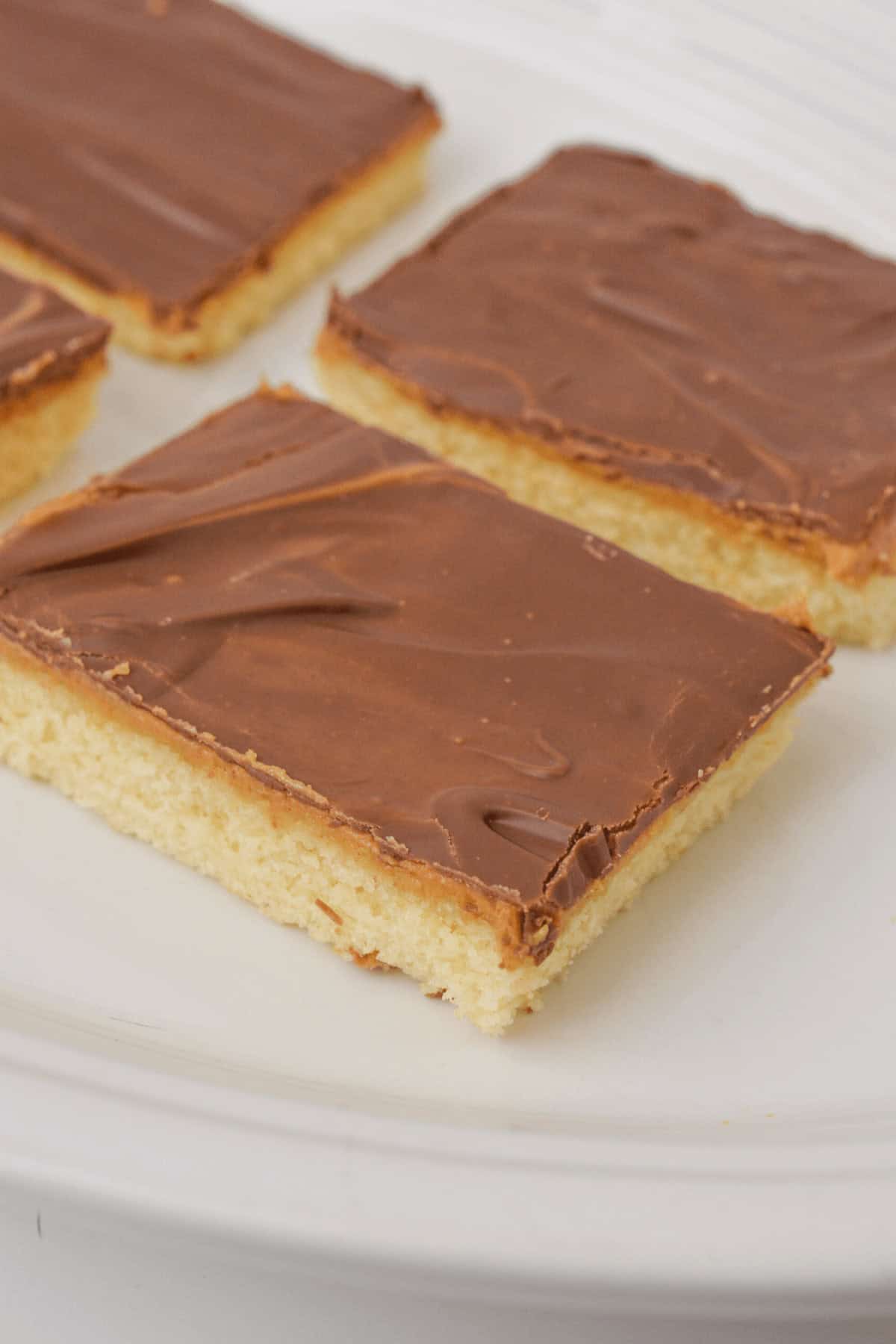 Four rectangular chocolate and peanut butter bars rest elegantly on a white plate, their tops glistening with a rich layer of chocolate.