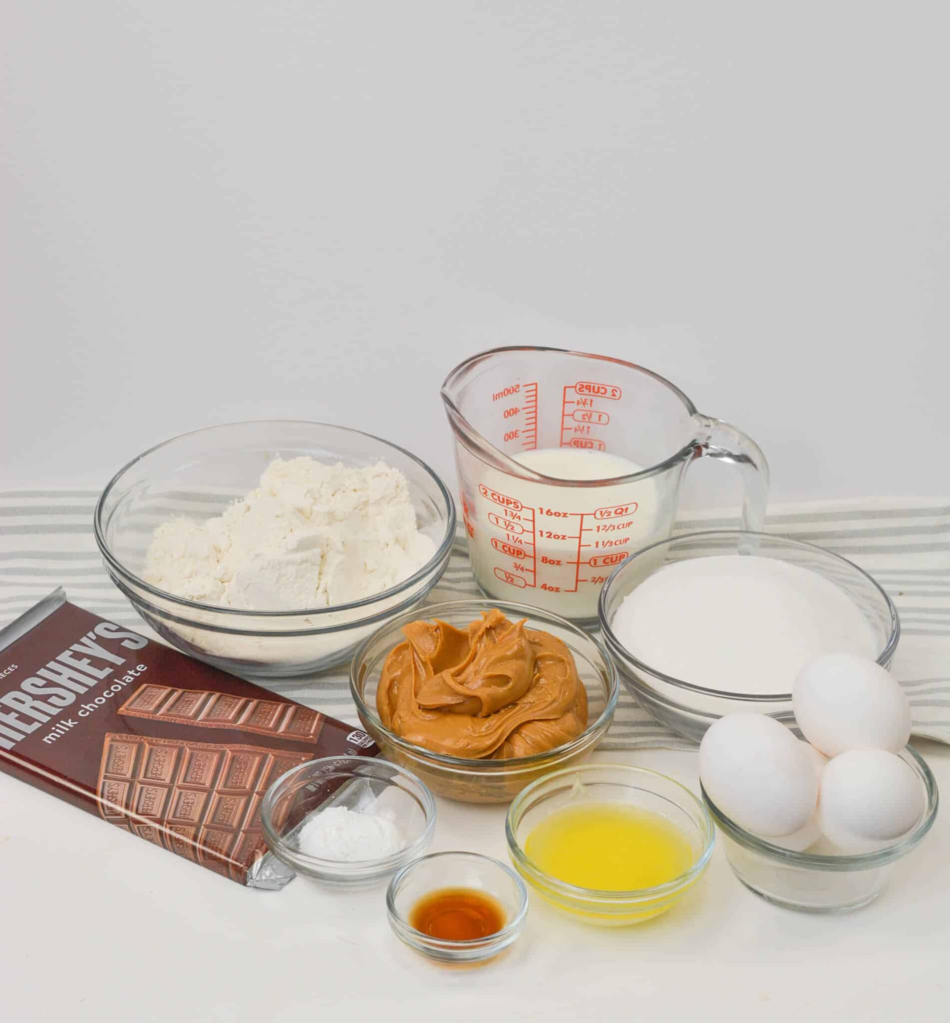 A collection of baking ingredients for a chocolate and peanut butter cake, featuring flour, milk, sugar, eggs, melted butter, vanilla extract, and baking powder on a striped cloth.