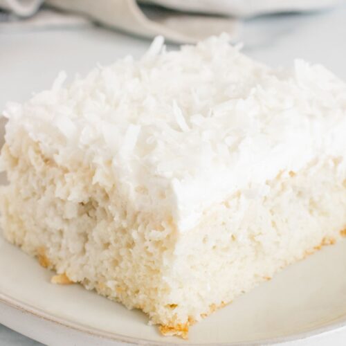 A square slice of Coconut Poke Cake with white frosting and shredded coconut on top, served on a light-colored plate.