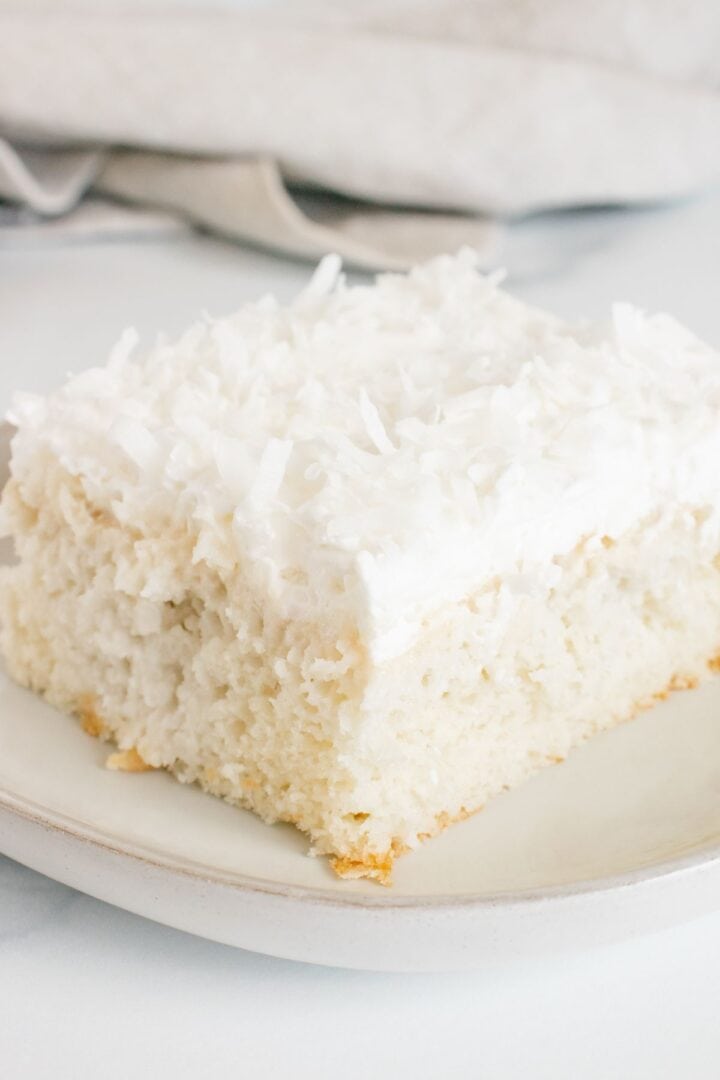 A square slice of Coconut Poke Cake with white frosting and shredded coconut on top, served on a light-colored plate.