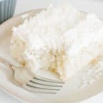 A square slice of Coconut Poke Cake topped with shredded coconut sits on a plate with a fork; a bite has been taken from the cake.