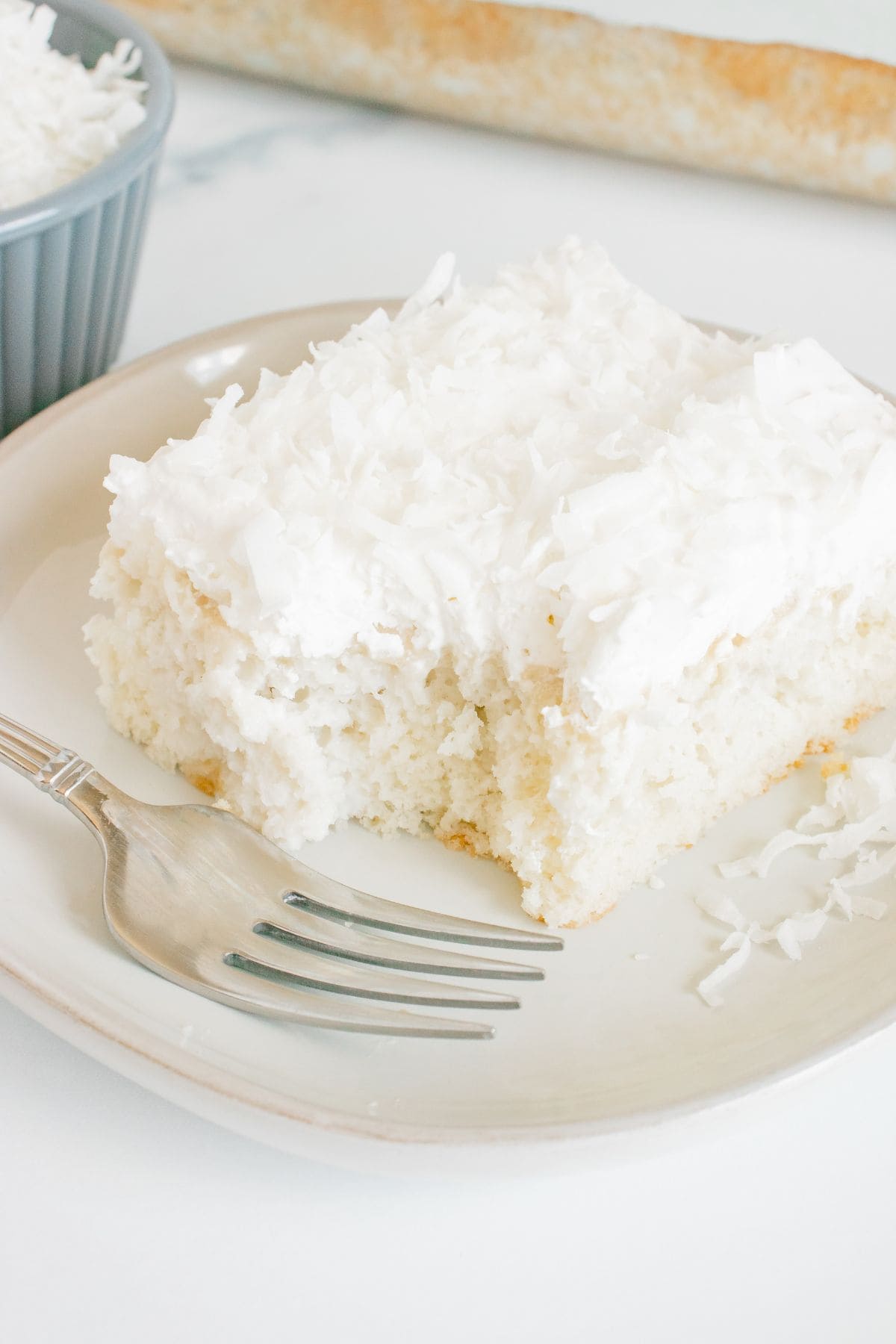 A square slice of Coconut Poke Cake topped with shredded coconut sits on a plate with a fork; a bite has been taken from the cake.