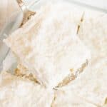 A square topped with shredded coconut, is being lifted from a glass baking dish filled with several other delicious cut pieces.