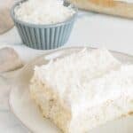 A slice of Coconut Poke Cake with white frosting and shredded coconut sits on a plate, accompanied by a small bowl of shredded coconut and a baking dish in the background.