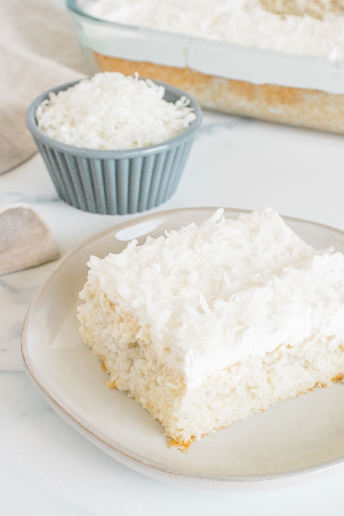 A slice of Coconut Poke Cake with white frosting and shredded coconut sits on a plate, accompanied by a small bowl of shredded coconut and a baking dish in the background.
