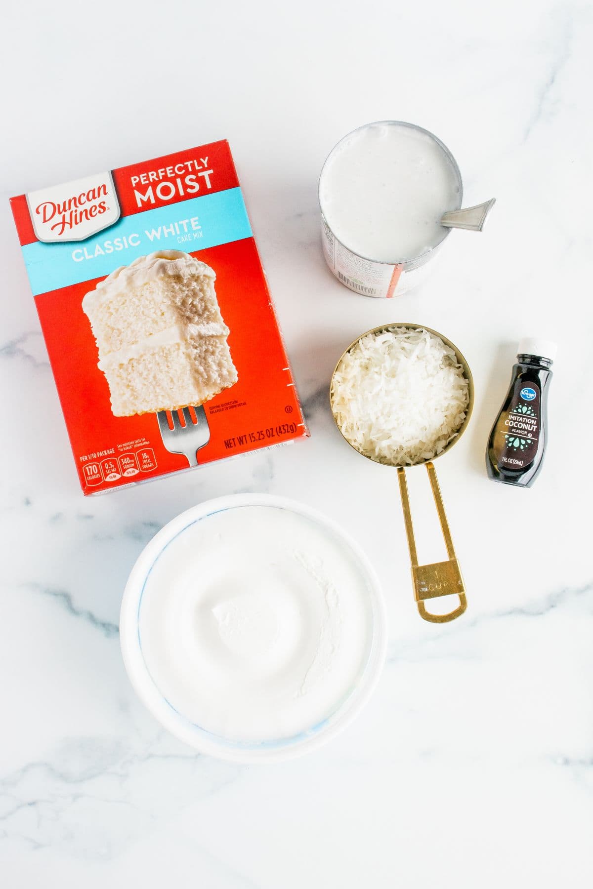 A box of Duncan Hines cake mix, milk in a measuring cup, extract, and a bowl of white powder—all the essentials to make a delicious dessert—are arranged on a white surface.