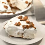 A slice of cream pie with chocolate pieces and a hint of caramel delight sits on a plate, next to the remaining pie in a pie tin.