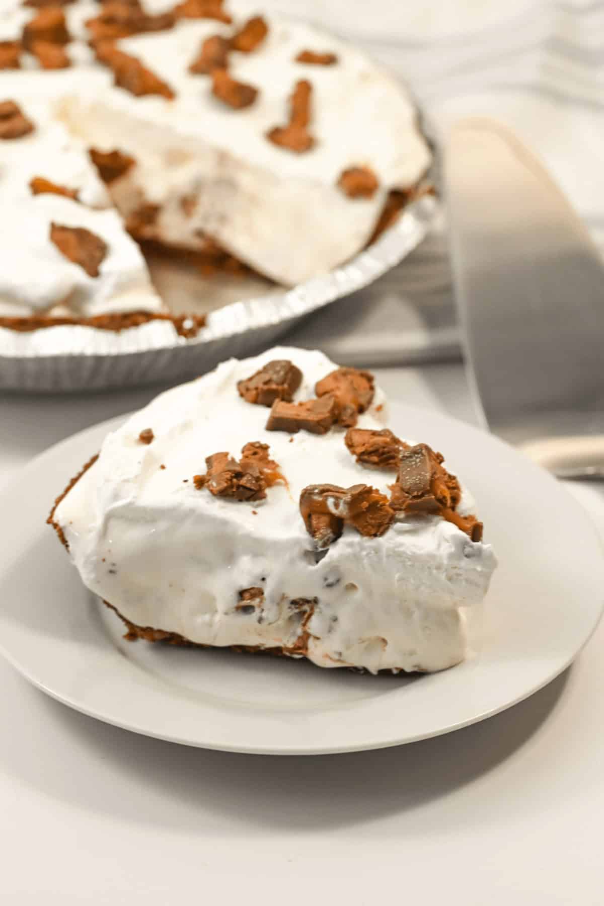A slice of cream pie with chocolate pieces and a hint of caramel delight sits on a plate, next to the remaining pie in a pie tin.
