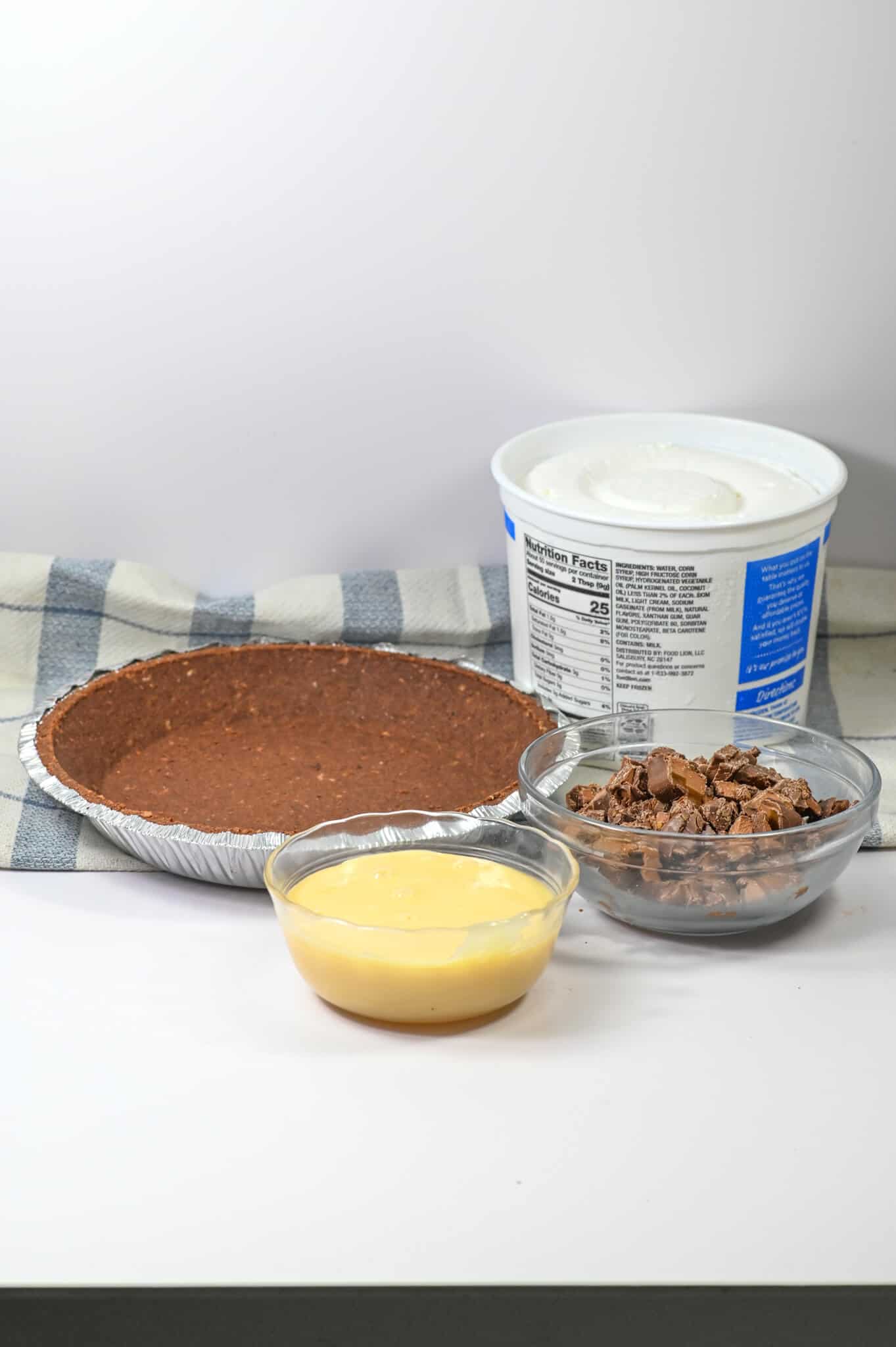 Ingredients for a caramel pie: an empty chocolate crust, a bowl of yellow pudding, a bowl of crumbled chocolate, and a tub of whipped topping, all on a checkered cloth.